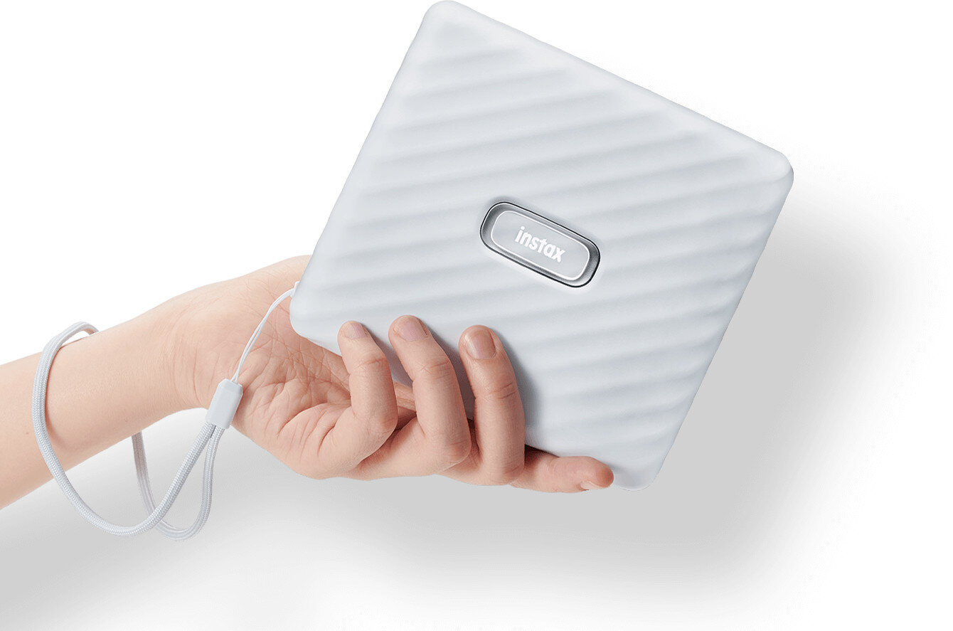 Biały aparat Instax w ręce osoby z paskiem na nadgarstek. Białe tło.