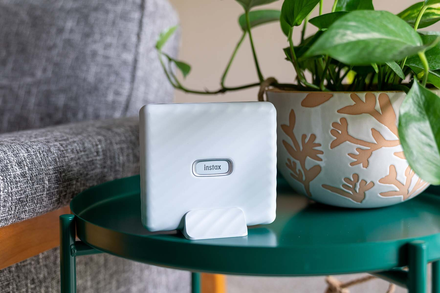 Biała drukarka Instax stoi na zielonym stoliku obok rośliny doniczkowej na szarym krześle.