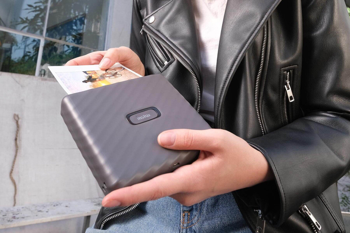 Ręce trzymające szarą drukarkę Instax i wydrukowane zdjęcie, z kurtką skórzaną.