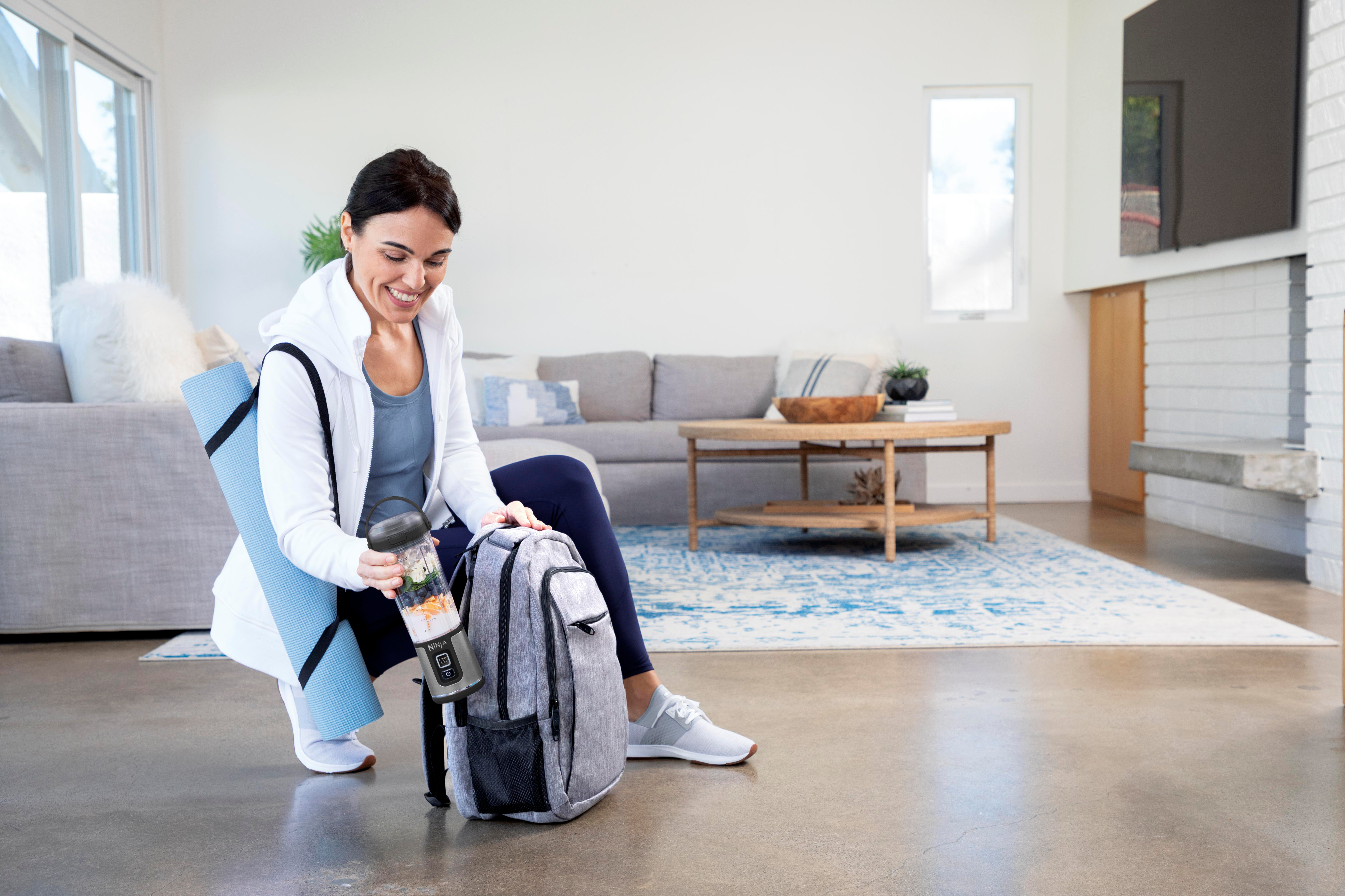 Frau mit Mixer, Yogamatte und Rucksack. Lächelnd. Hintergrund umfasst Couch, Teppich und Couchtisch.
