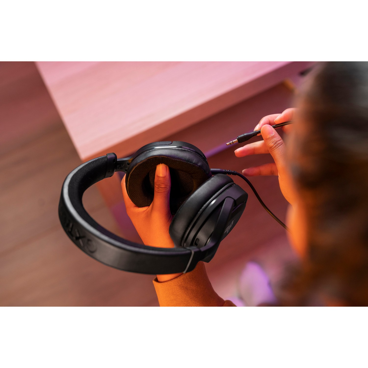 A person connects a black gaming headset to a device on a wooden table.