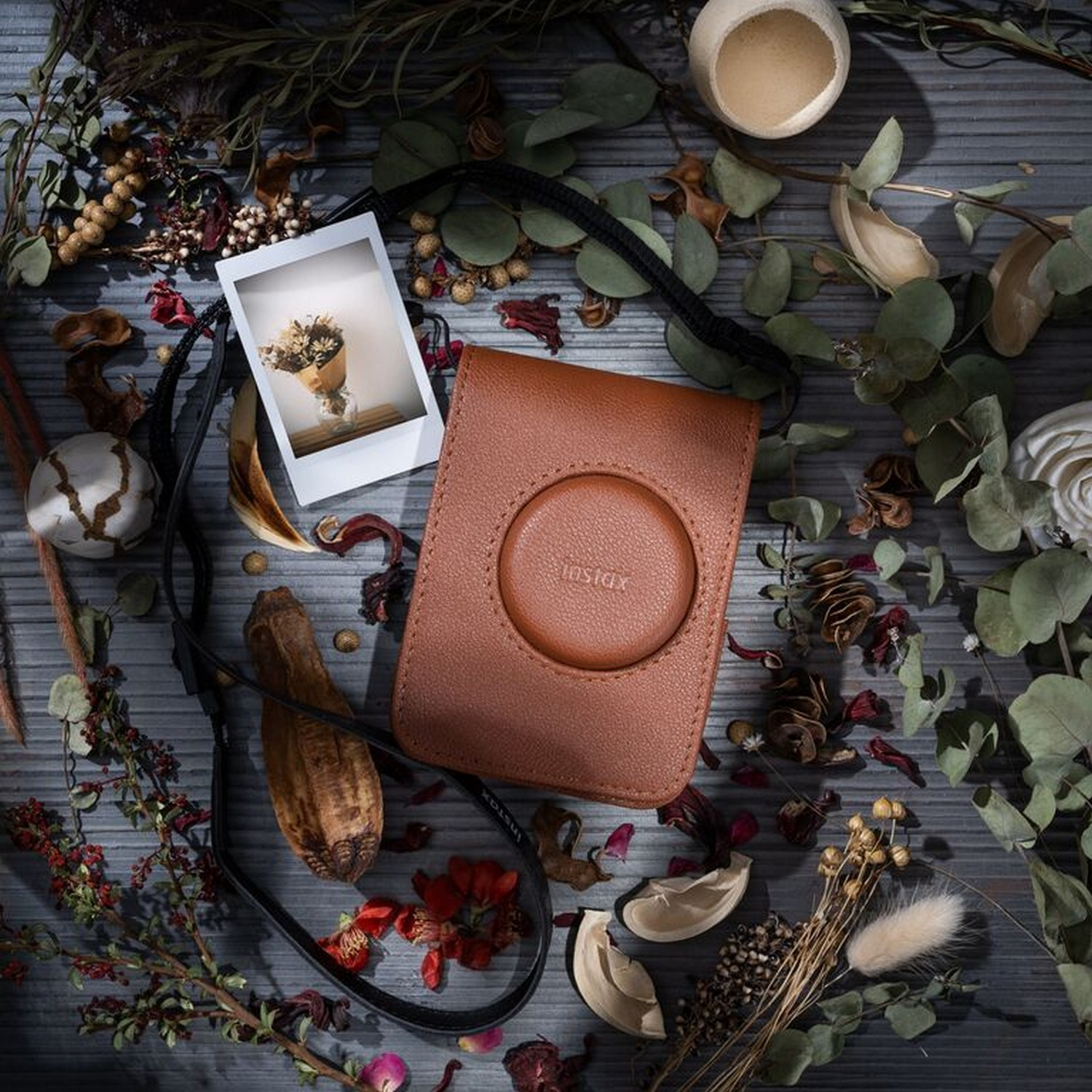 Polaroid photo, Instax camera case, dried flowers on wooden surface.