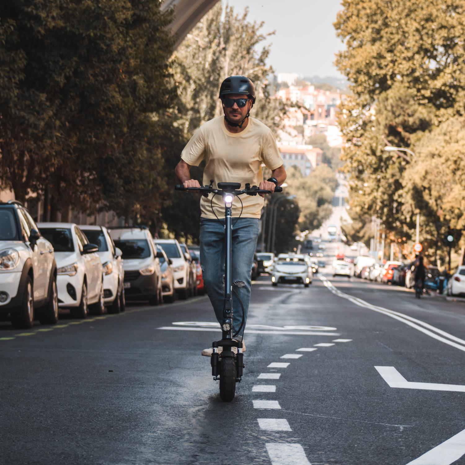 Un hombre monta un patinete eléctrico por una calle. Lleva casco y gafas de sol. Coches a los lados.