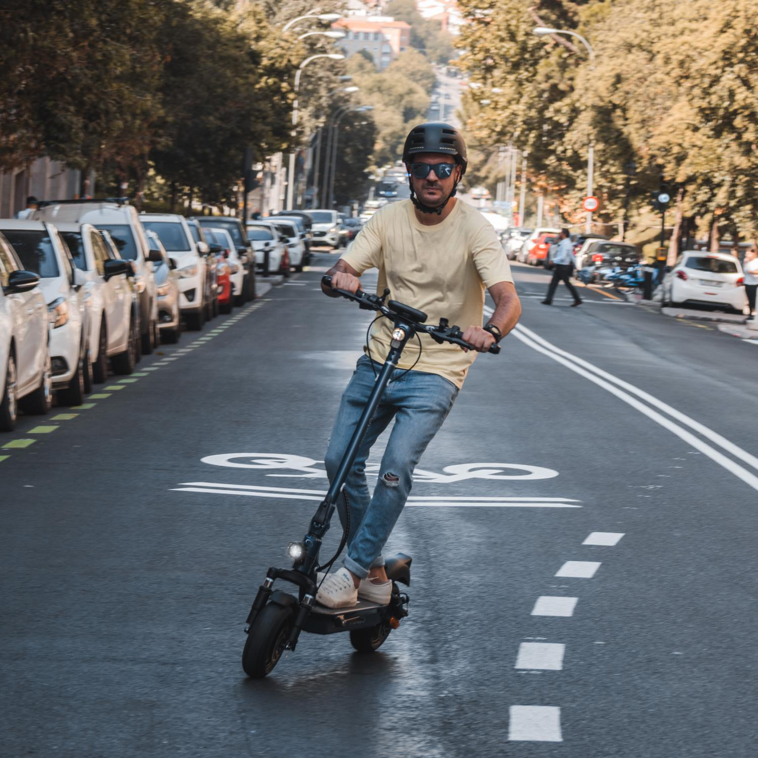 Hombre montando un patinete eléctrico en una carretera. Con casco y gafas de sol, coches a los lados.