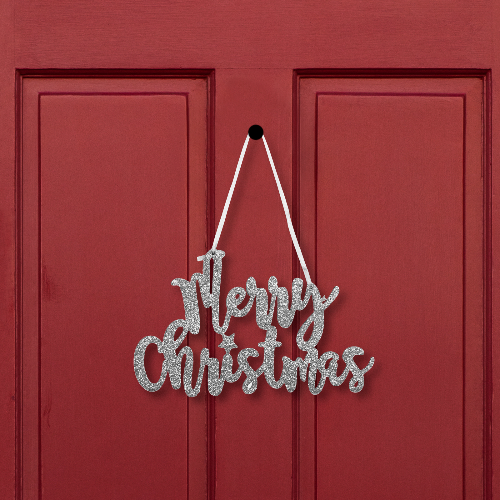 Egy ezüst csillogó "Merry Christmas" felirat lóg egy piros ajtón, fehér szalaggal rögzítve.