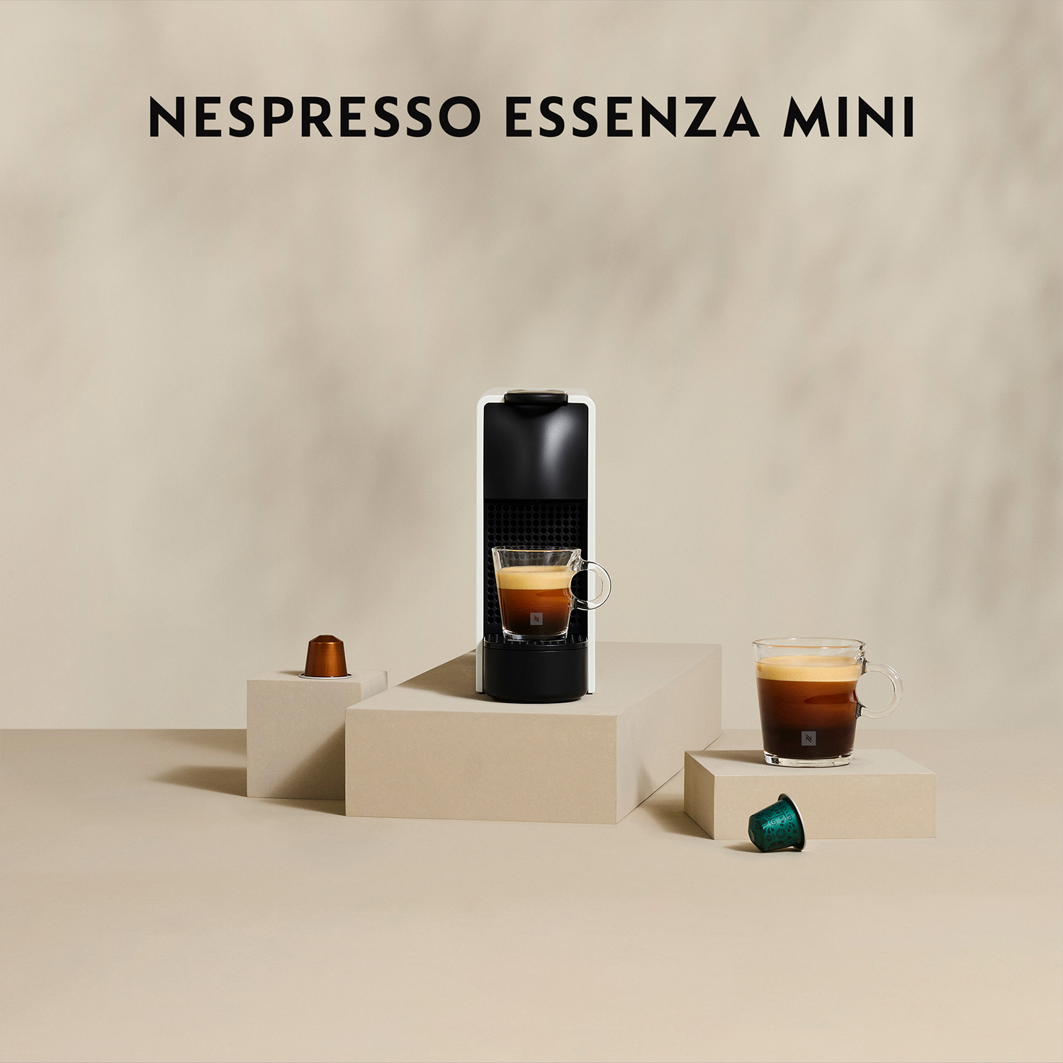 Macchina da caffè Nespresso Essenza Mini. Due tazze di caffè e una capsula. Sfondo neutro.