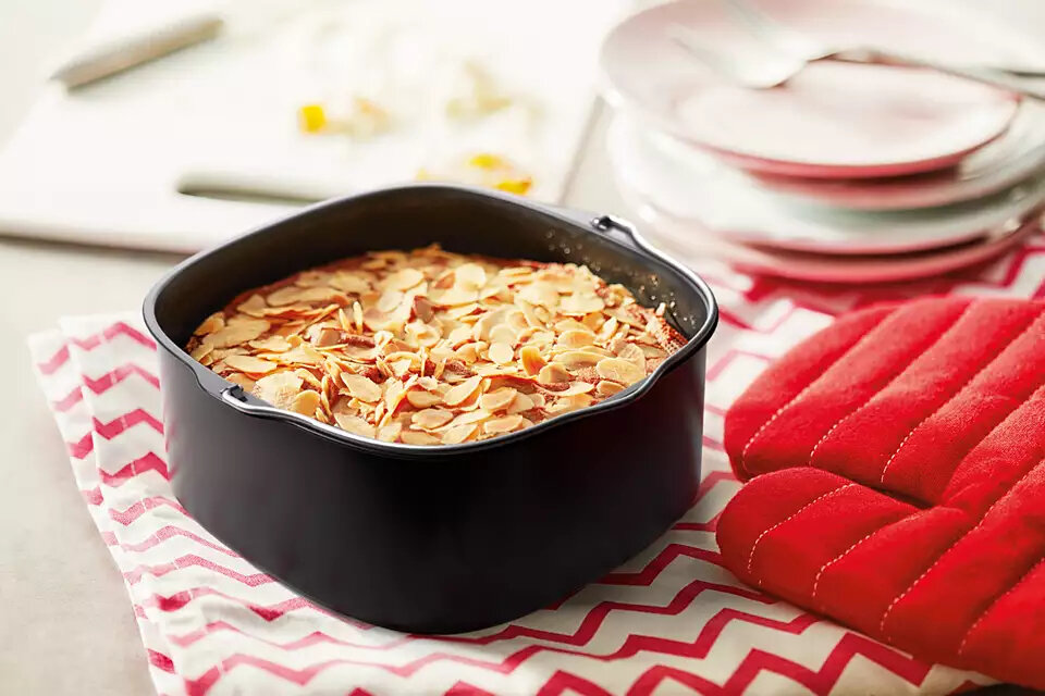 Ciasto w czarnej formie do pieczenia ze płatkami migdałów, z czerwonymi rękawicami kuchennymi.