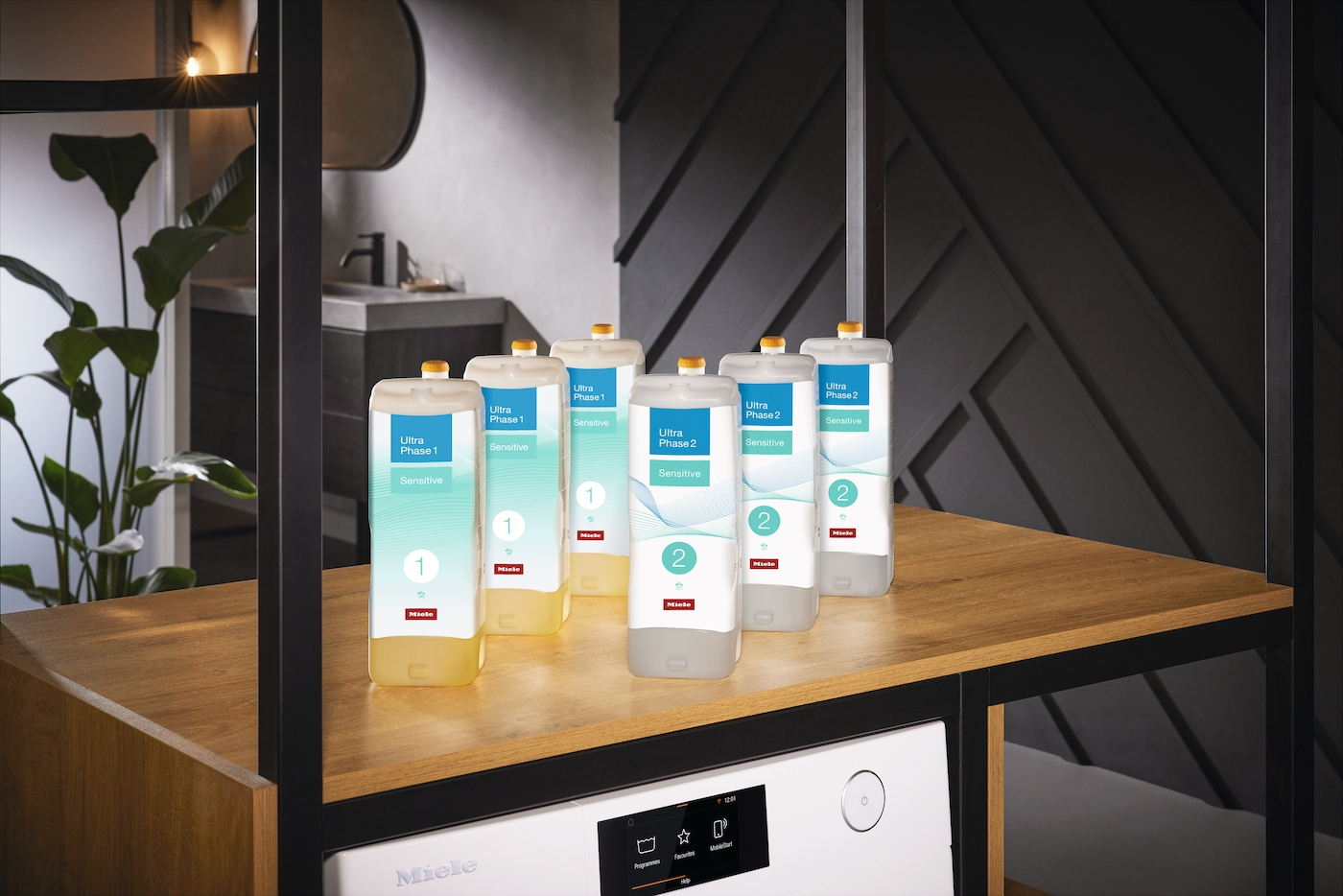 Sechs Miele-Waschmittelflaschen mit der Aufschrift Ultra Phase 1 und 2, Sensitive. Sie stehen auf einem Holzregal, darunter eine Waschmaschine.