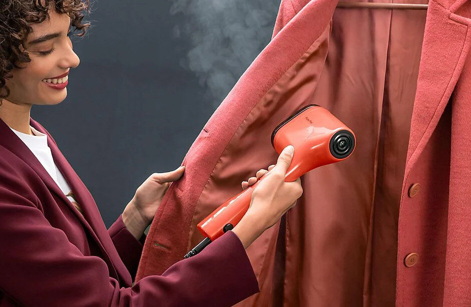 Kobieta prasuje czerwony płaszcz pomarańczowym parownikiem. Uśmiecha się.