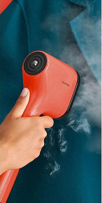 Ręka trzymająca czerwoną parownicę do ubrań nad niebieskim ubraniem, widoczna para.