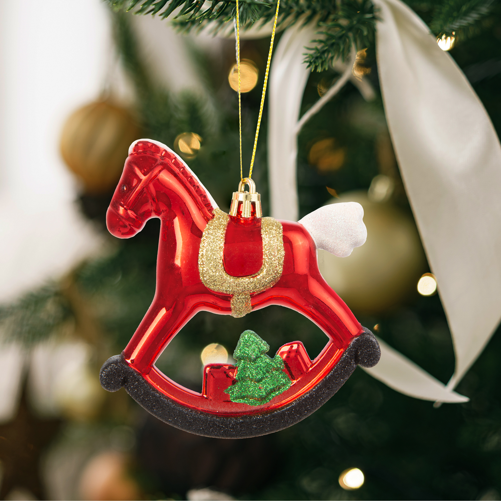 Egy piros hintaló karácsonyi dísz arany csillámos díszítéssel, zöld fával és fehér szárnyakkal, ami a fán lóg.