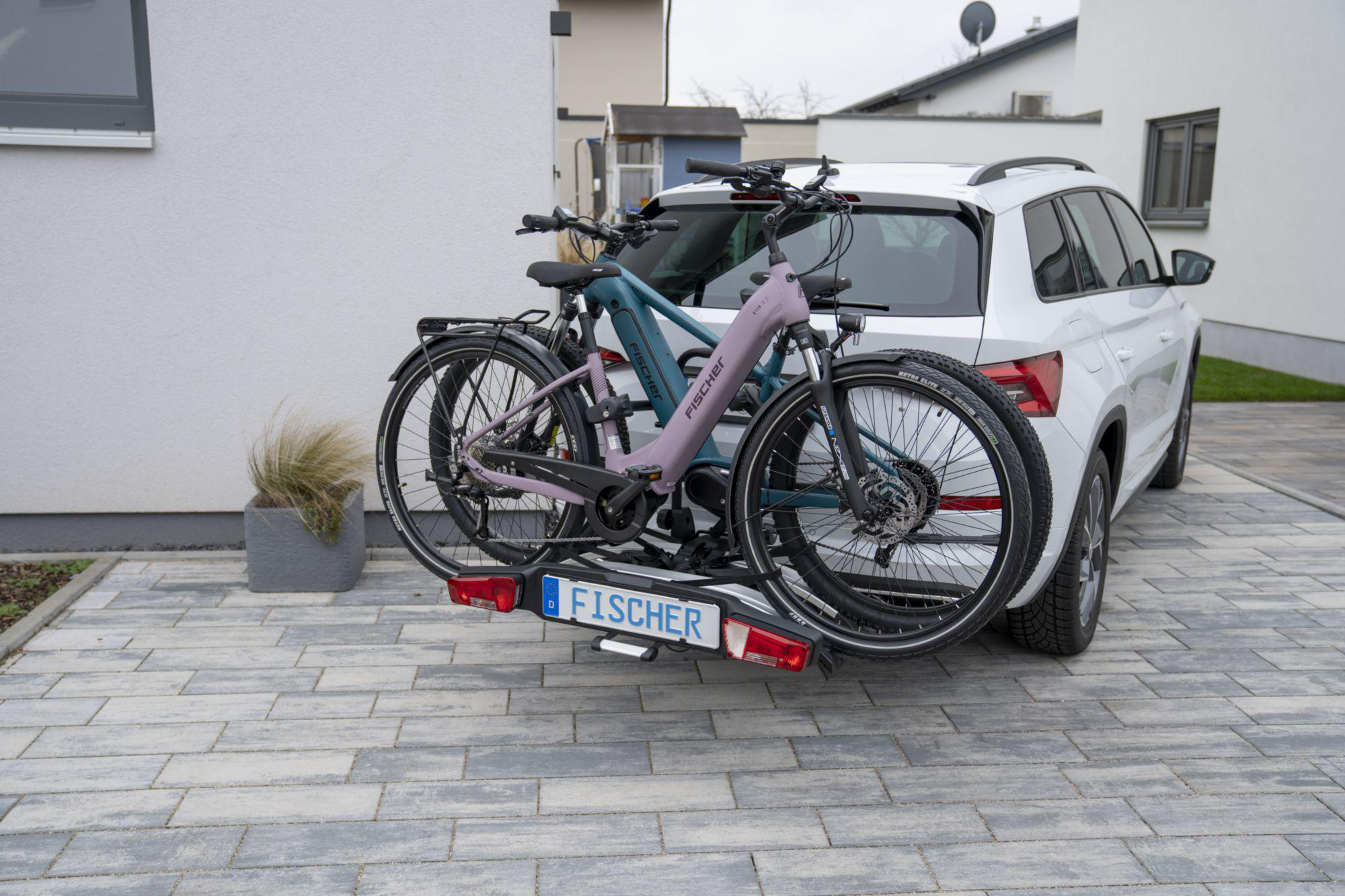 Zwei Fahrräder auf einem Autoträger, geparkt auf einer Backstein-Auffahrt. Weißes Auto im Hintergrund.