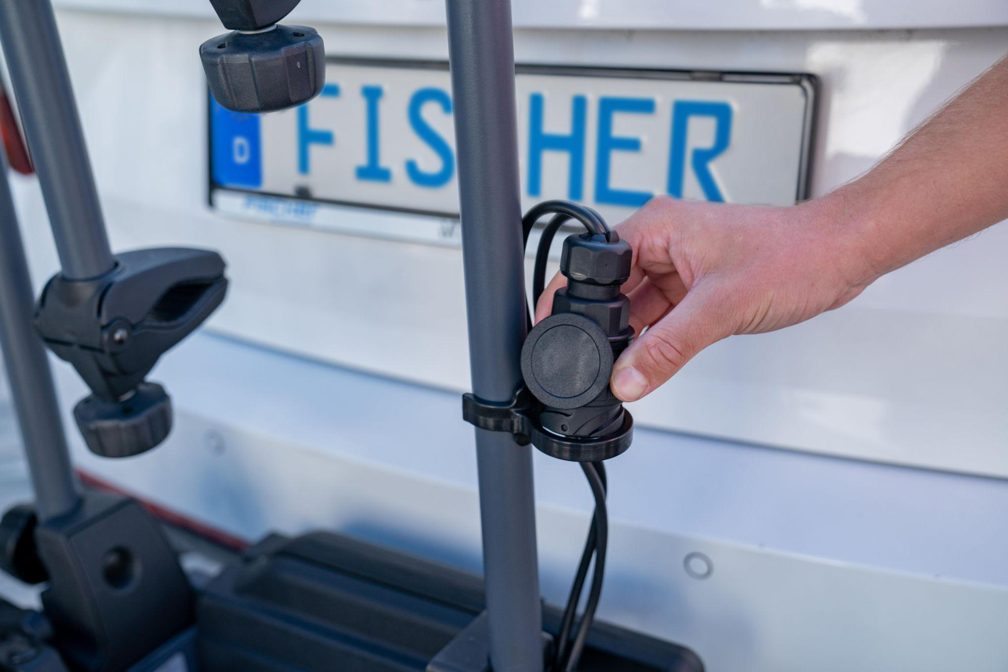 Die Hand einer Person verbindet einen schwarzen Stecker mit einem Fahrradträger eines Autos, mit Kennzeichen.