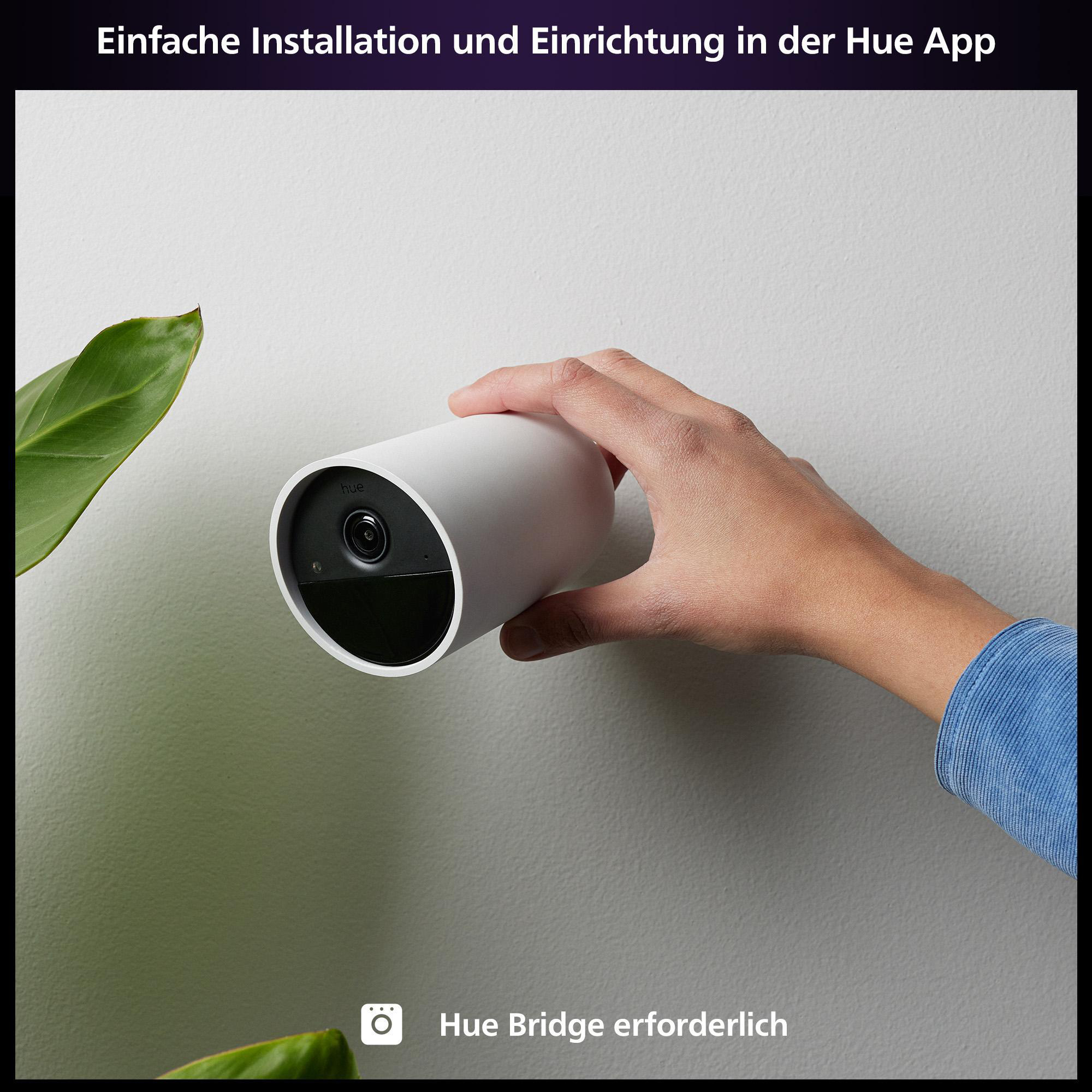 Hand hält weiße Smart-Kamera an weißer Wand, Pflanze sichtbar.