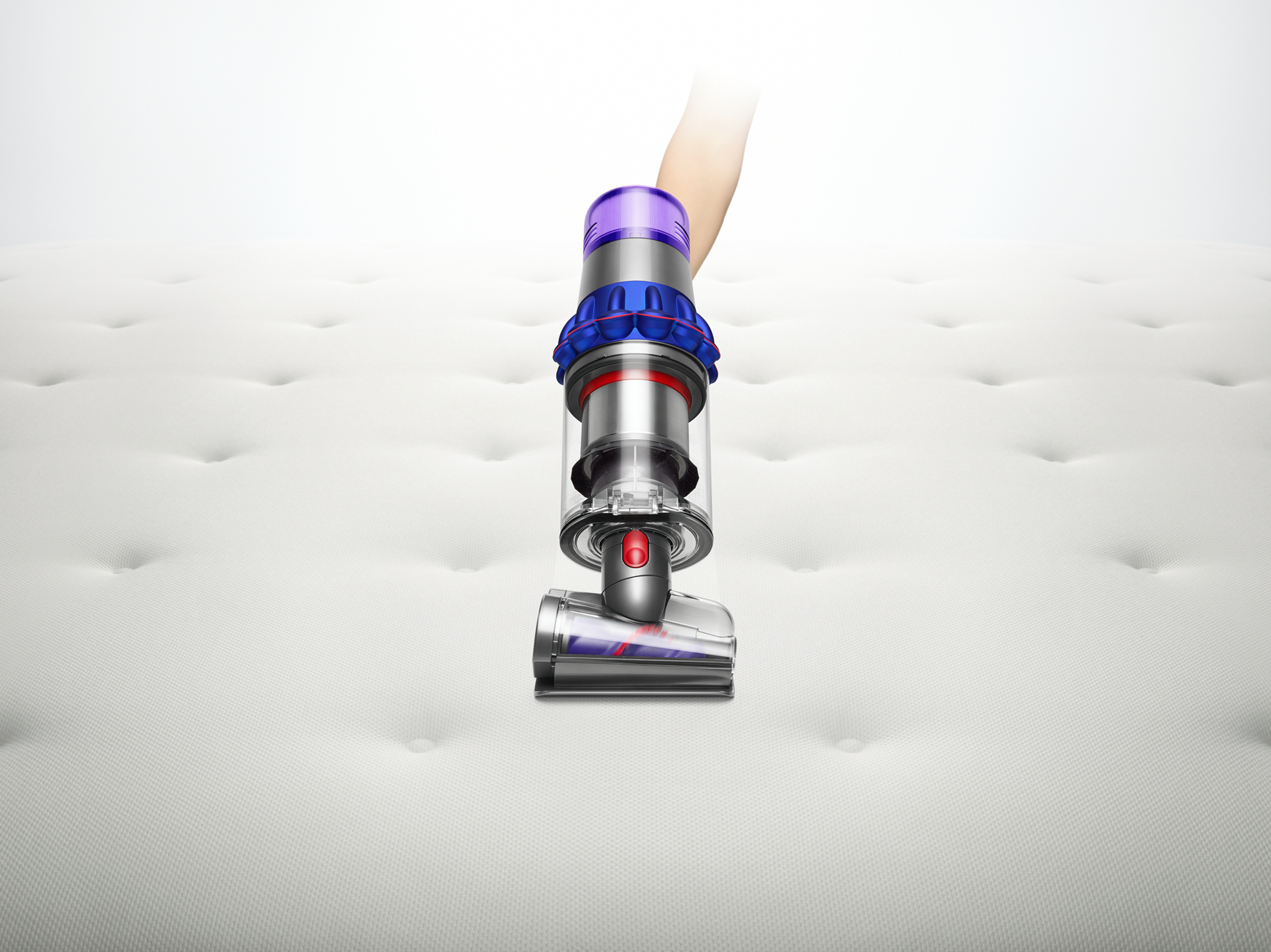 Una aspiradora Dyson en un colchón blanco. Una mano sostiene el dispositivo morado y azul.