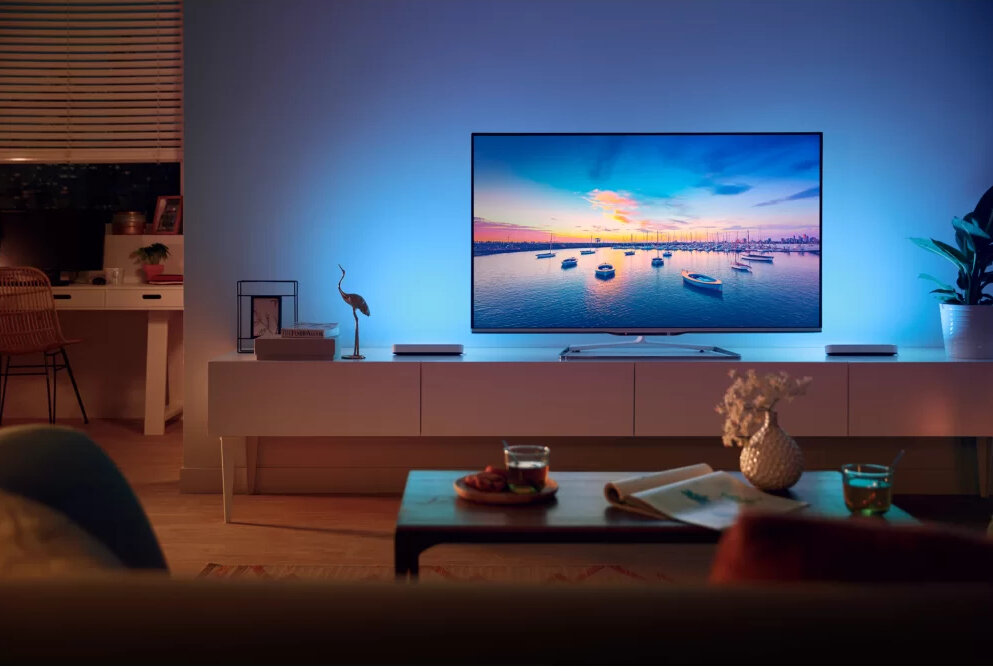 Telewizor zamontowany na białej szafce. Telewizor pokazuje scenę zachodu słońca. Niebieskie światła za telewizorem.