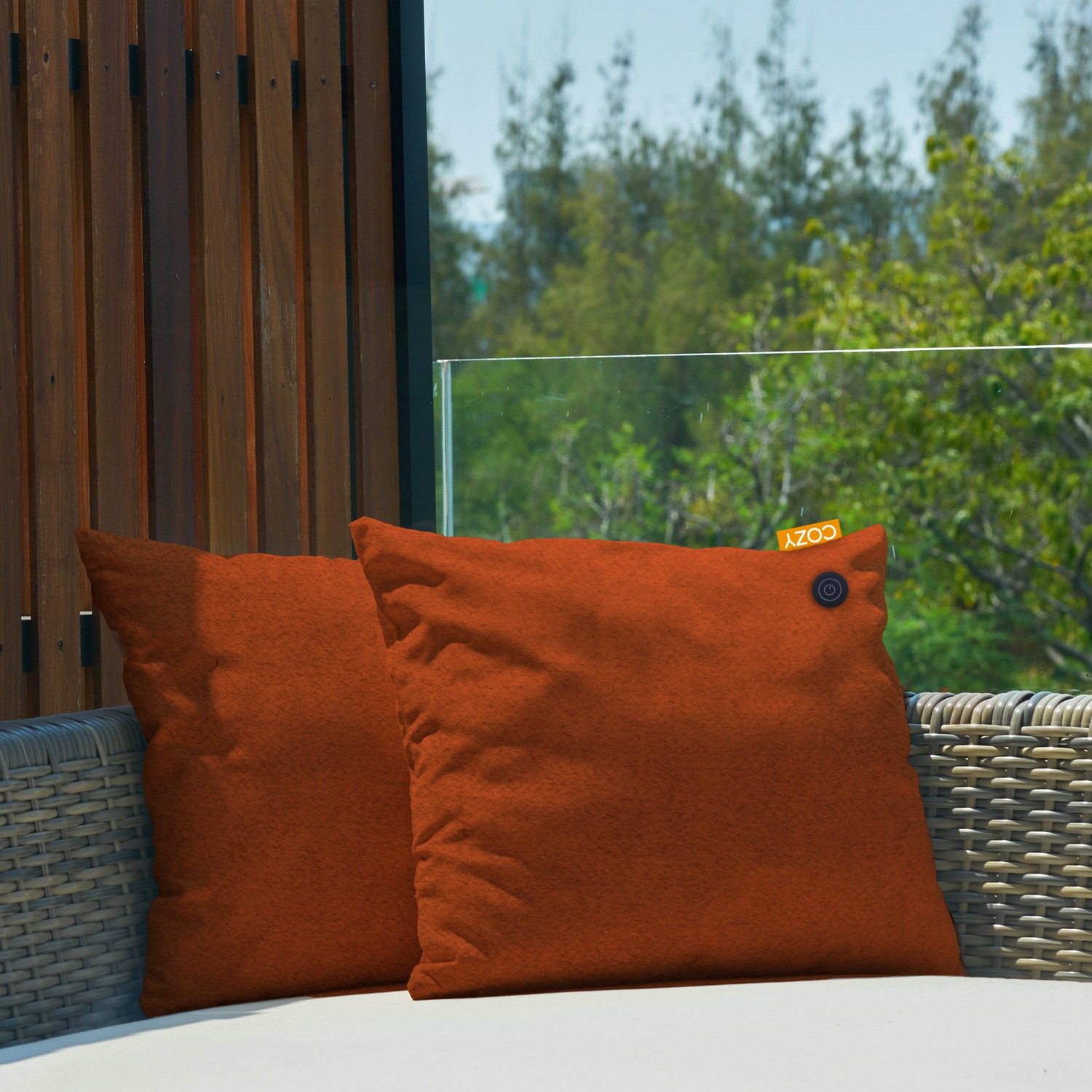 Deux coussins orange sur une surface blanche. Le fond montre un mur en bois, du verre et un arbre vert.