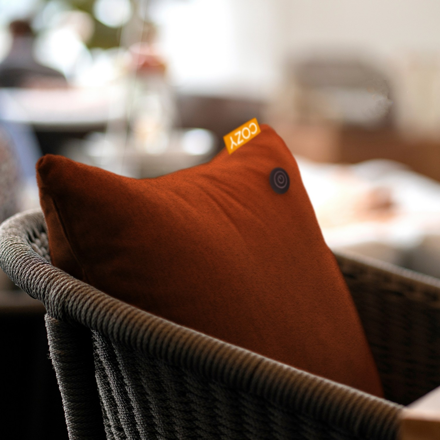 Un oreiller orange avec l'étiquette 'COZY' repose dans une chaise tissée. L'oreiller a un bouton. Fond doux d'une pièce.