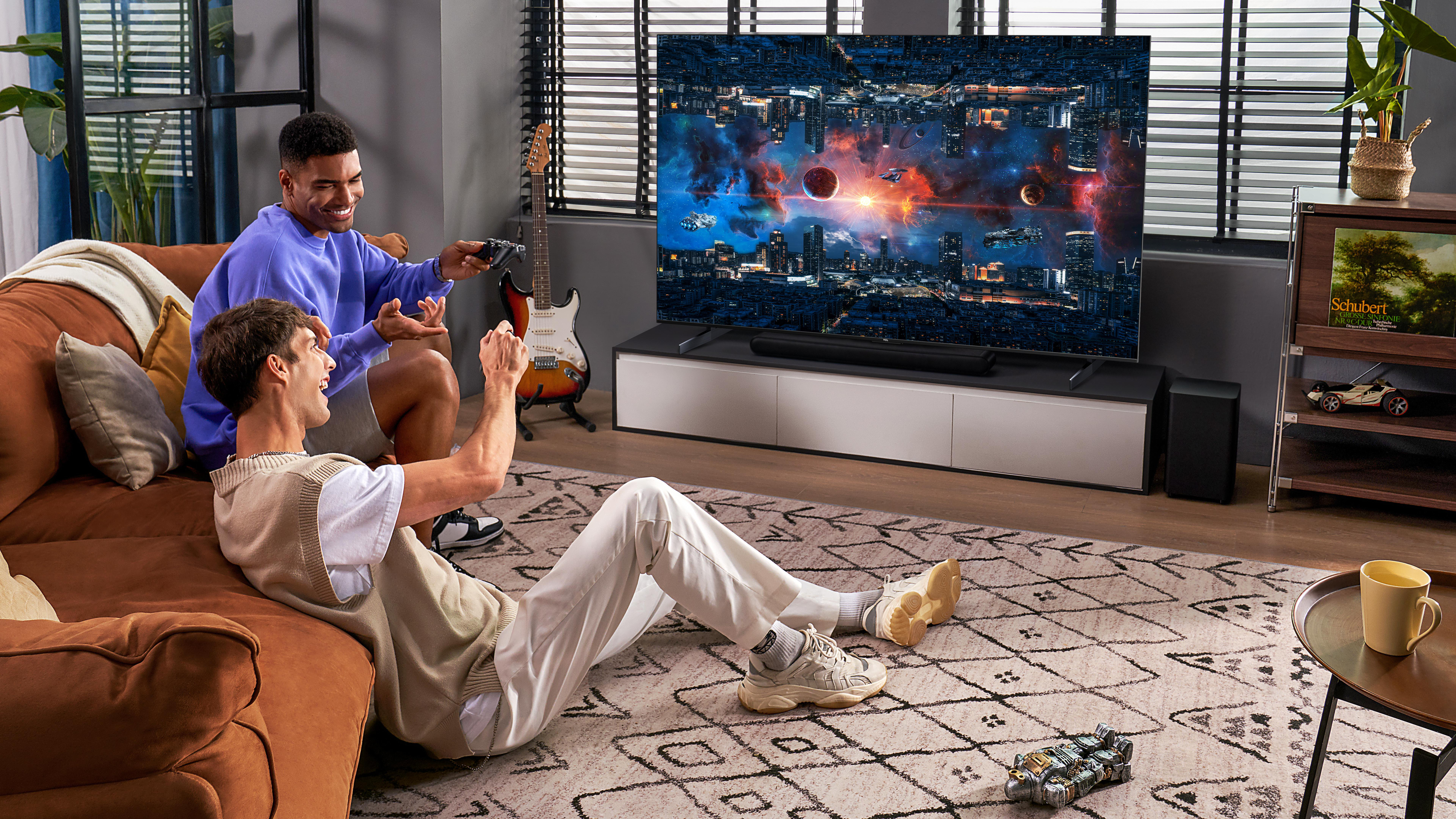 Zwei Männer spielen an einem großen Fernseher. Einer sitzt auf dem Sofa, der andere auf dem Boden.