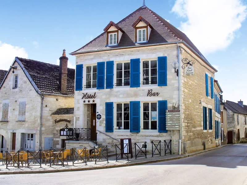 Un hôtel dans un village français, avec des volets bleus, un restaurant et un panneau indiquant le nom de la ville.