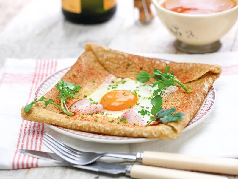 Una crepe con uovo, prosciutto e prezzemolo su un piatto, con una piccola ciotola e posate nelle vicinanze.
