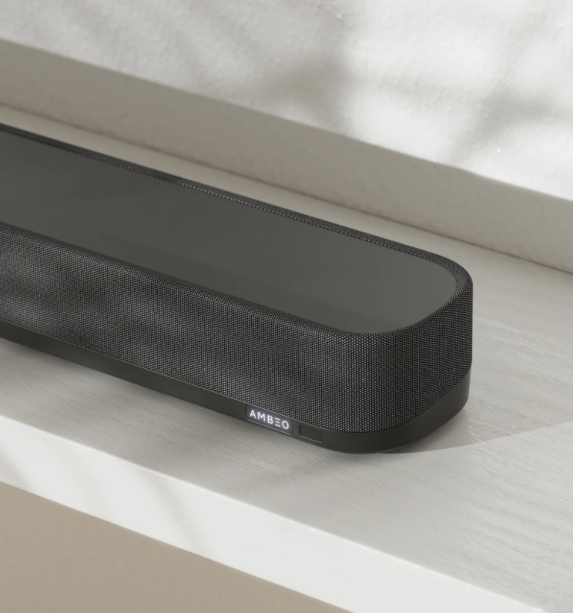 Fekete soundbar fehér polcon. Az 'AMBEO' szó látható az elején.