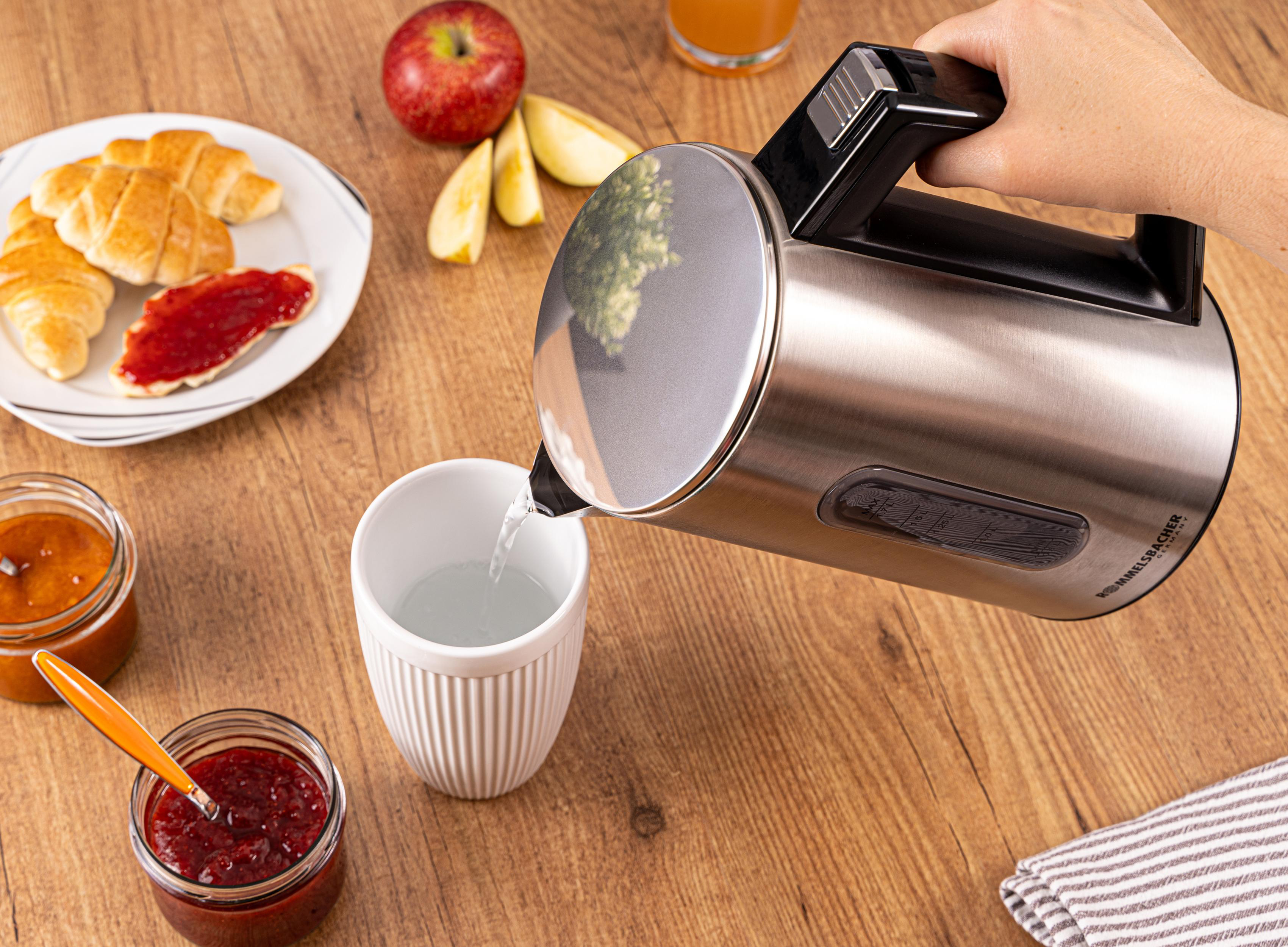 Person gießt Wasser aus silberner Kanne in weiße Tasse. Frühstücksartikel auf dem Tisch.