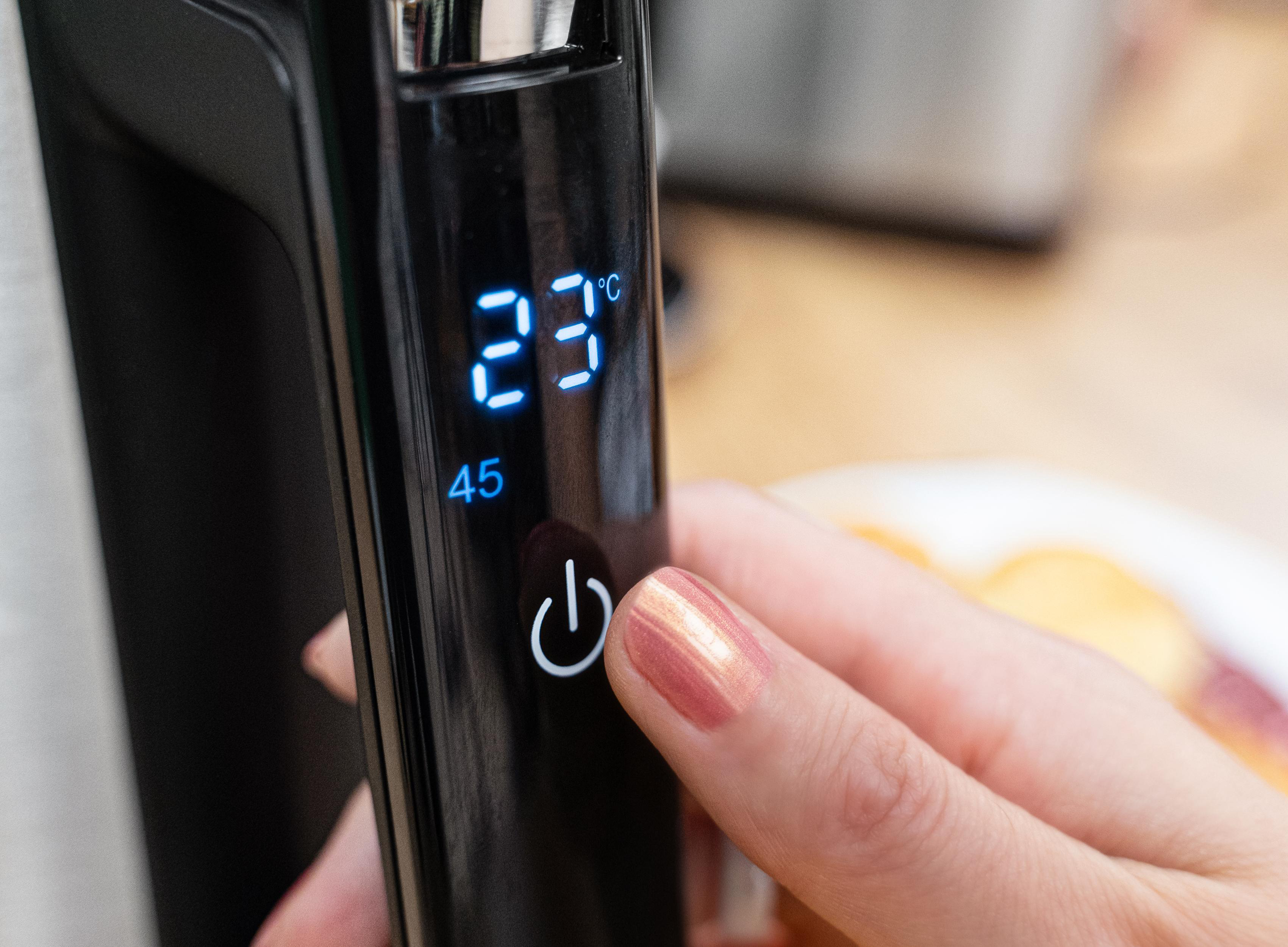 Hand drückt Knopf auf einem schwarzen Gerät mit 23°C. Im Hintergrund ein Teller mit Essen.