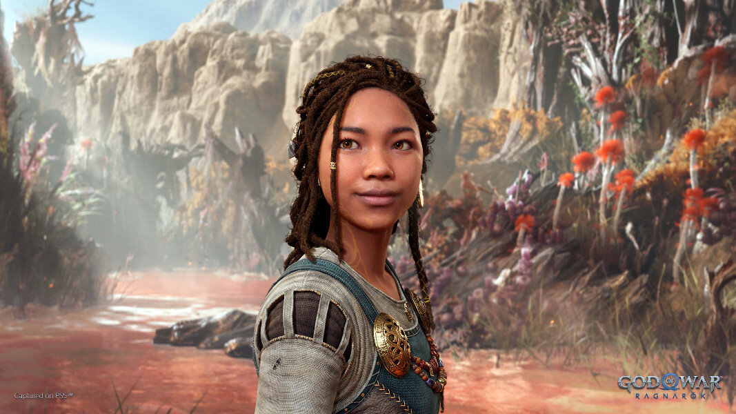 Kobieta z dredami, uśmiechnięta. Ma na sobie ozdobną biżuterię i niebiesko-kremowy top. Tło: skalisty krajobraz.