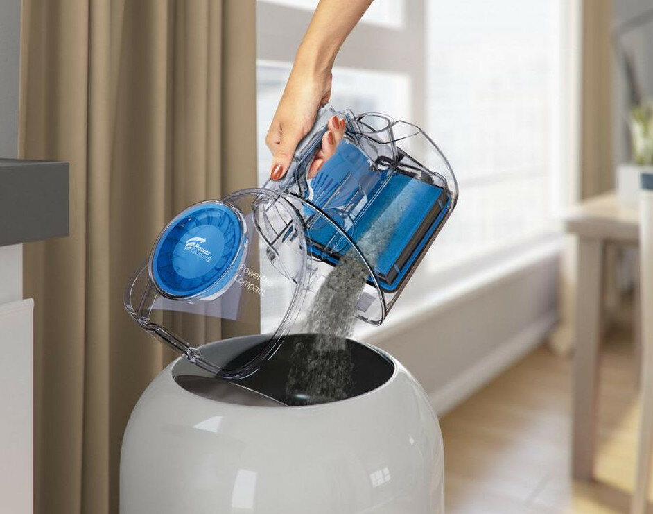 Kobieta opróżnia odkurzacz PowerCore Compact do białego kosza na śmieci. Widoczny jest kurz.