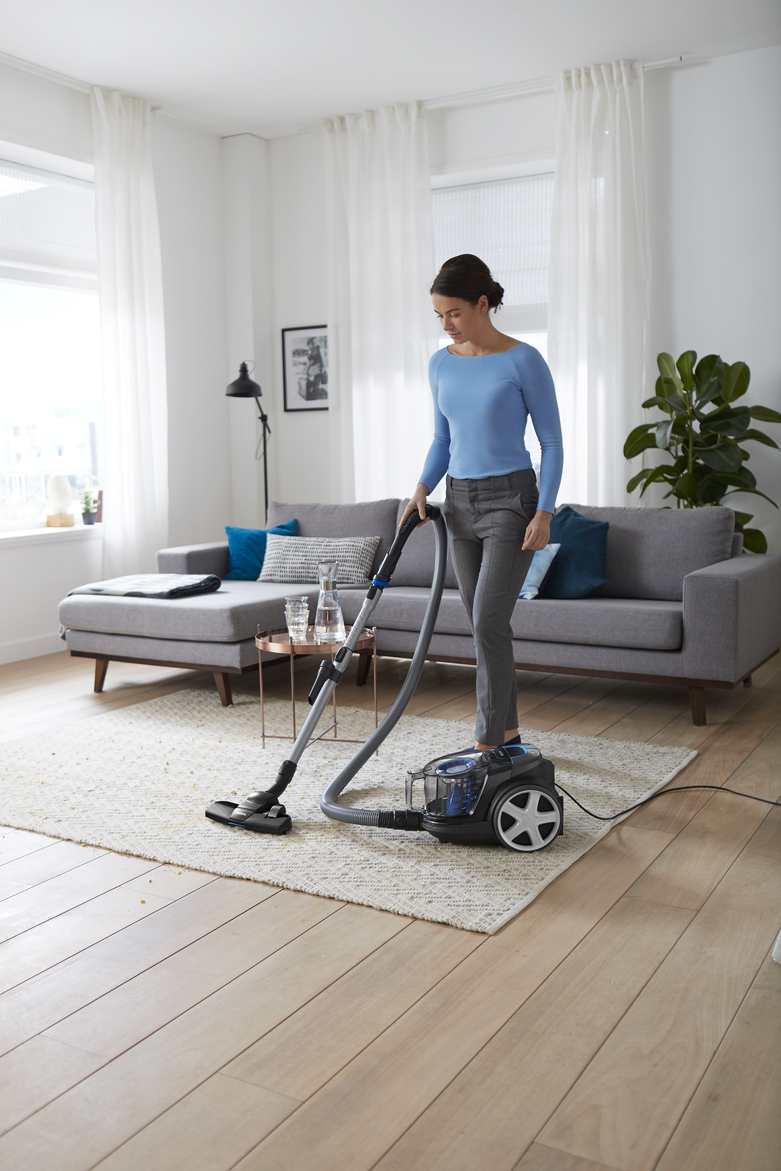 Kobieta odkurza salon z szarą sofą, dywanem i zasłonami. Obecna jest roślina i stół.