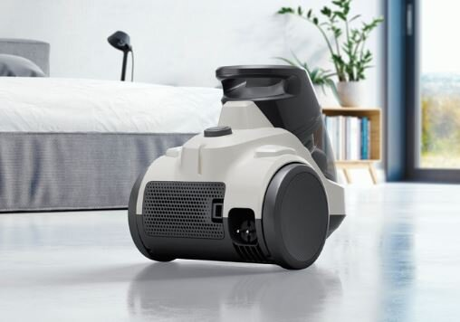 Biało-czarny odkurzacz na błyszczącej podłodze. W tle łóżko i roślina.