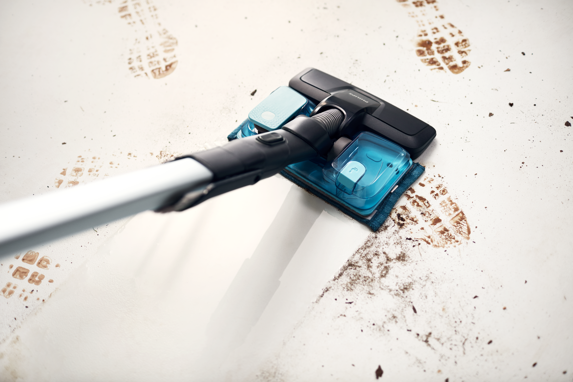Głowica odkurzacza czyszcząca podłogę. Widoczne zabrudzenia i ślady stóp. Widoczne niebieskie nakładki.