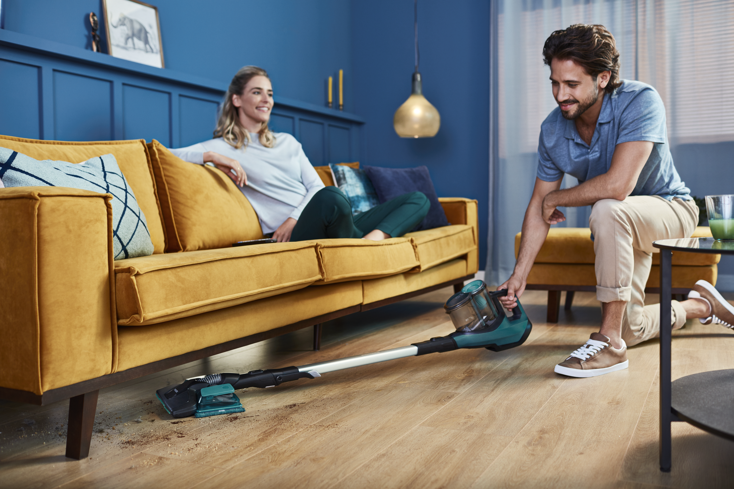 Mężczyzna odkurza drewnianą podłogę, a kobieta relaksuje się na żółtej sofie. Odpady na podłodze.
