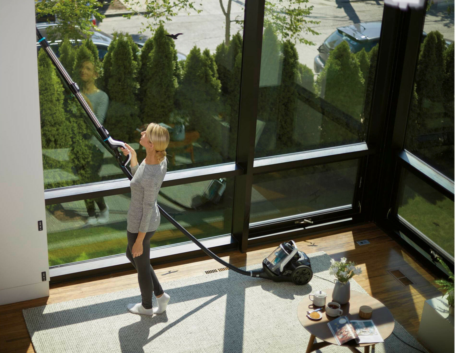Kobieta czyści okna odkurzaczem. Stół z przedmiotami na pierwszym planie.