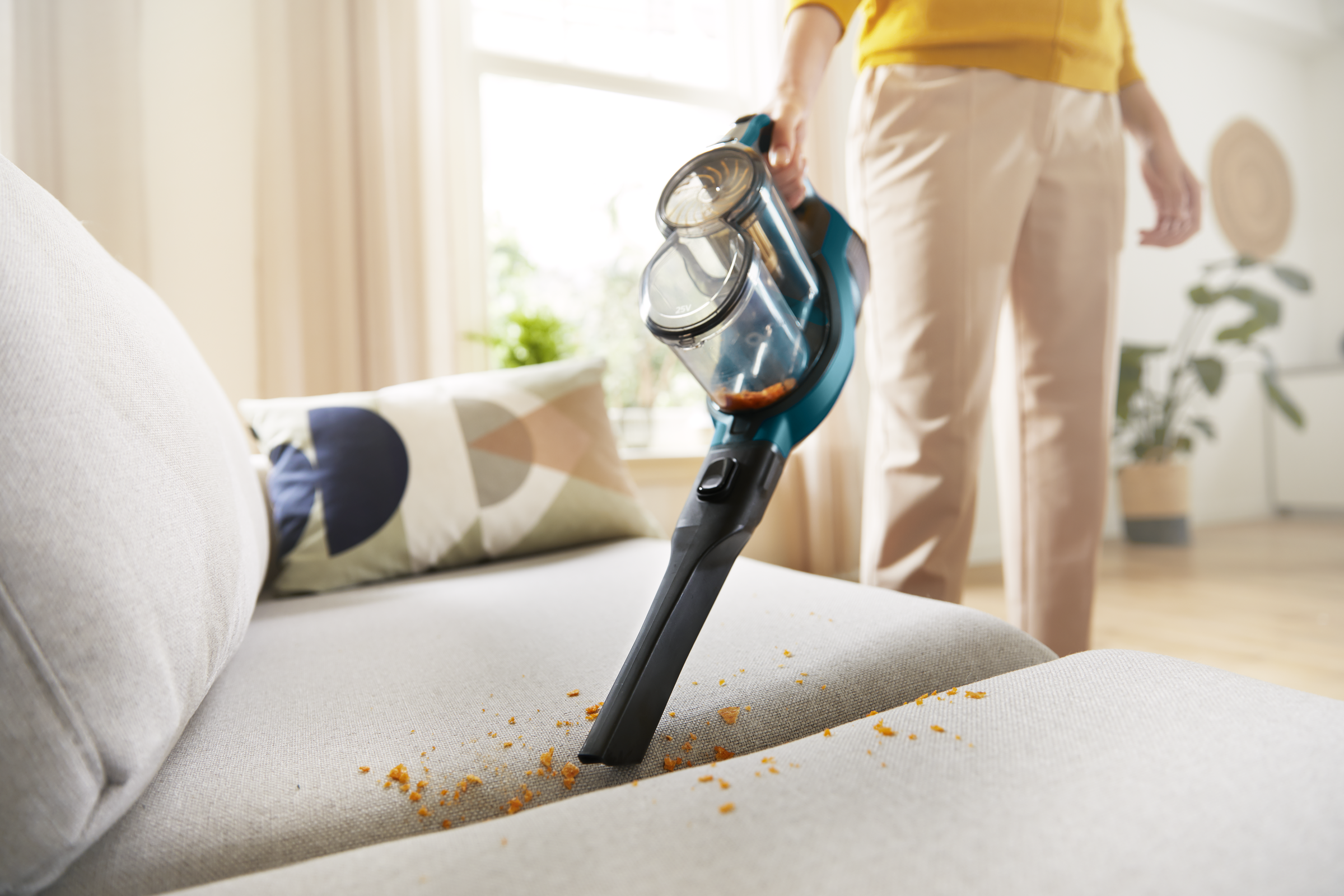 Osoba odkurzająca kanapę, odkurzacz jest skierowany w dół, usuwając okruchy. W tle widoczne okno.