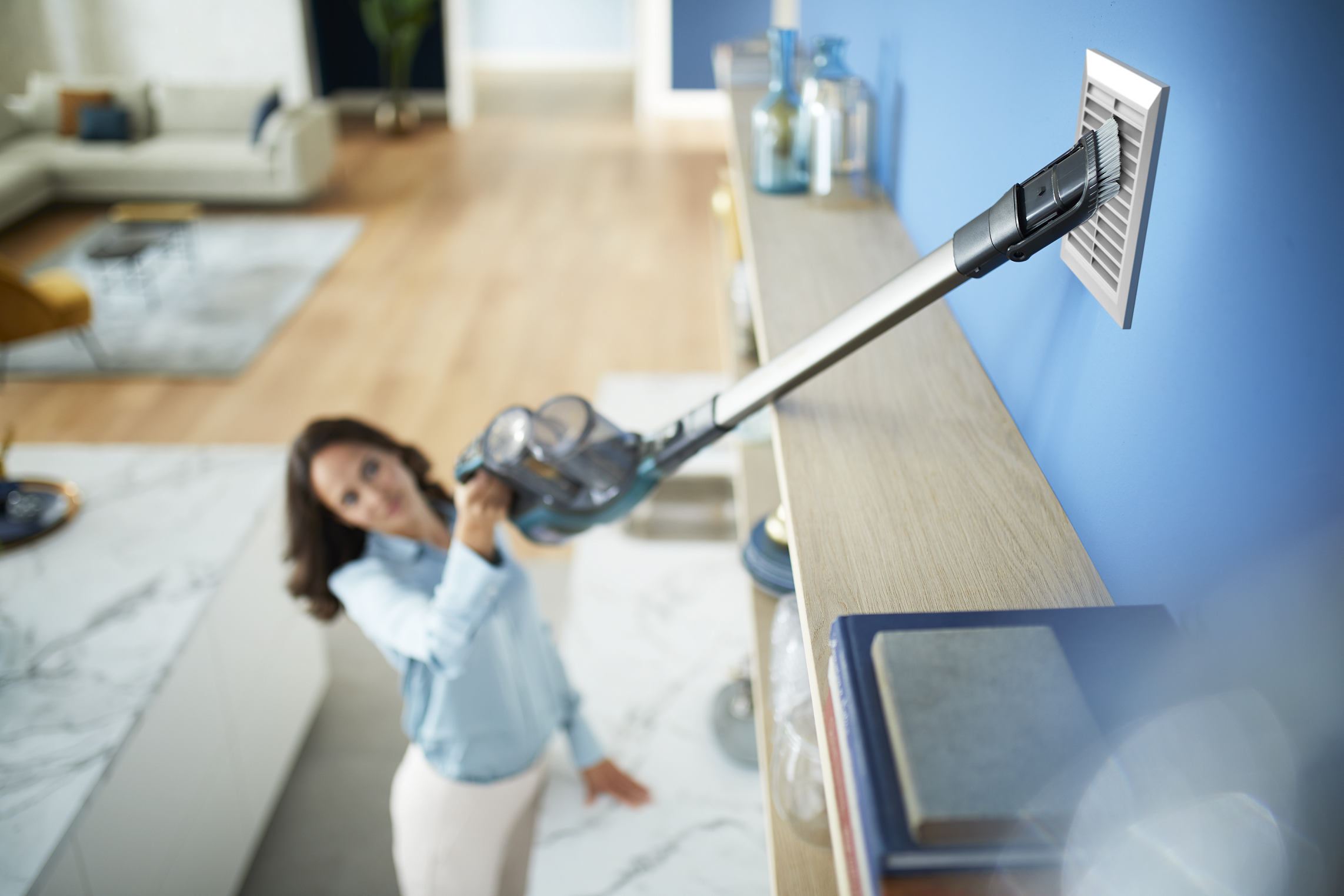 Kobieta odkurza kratkę wentylacyjną. Używa odkurzacza ręcznego z długą przedłużką.