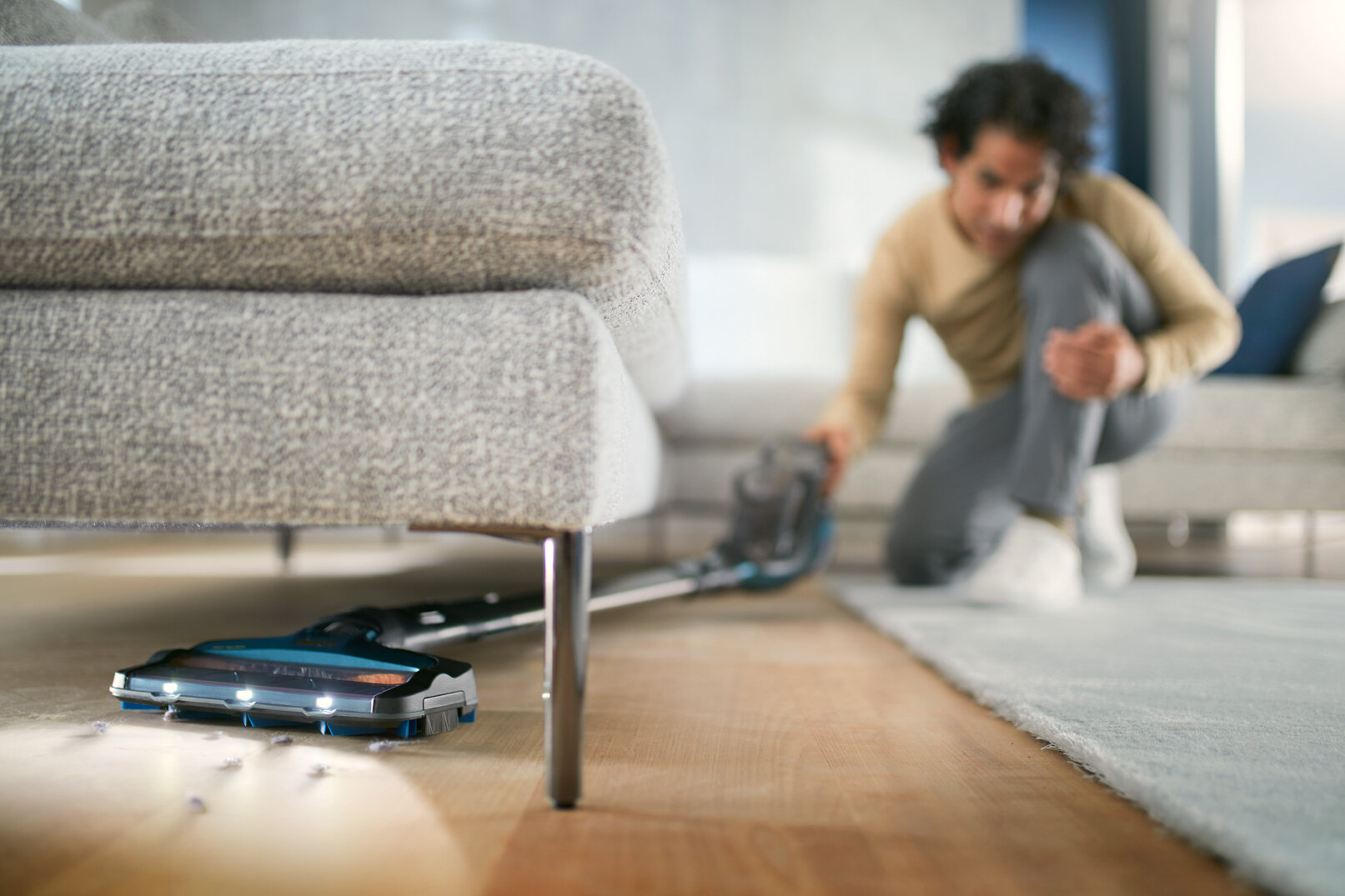 Osoba odkurzająca podłogę z szarą kanapą i dywanem w tle.