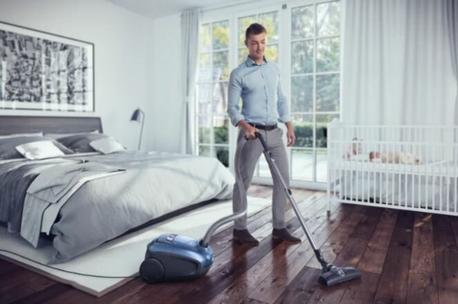 Mężczyzna odkurza sypialnię. W tle łóżko, kołyska i ramka na zdjęcia.