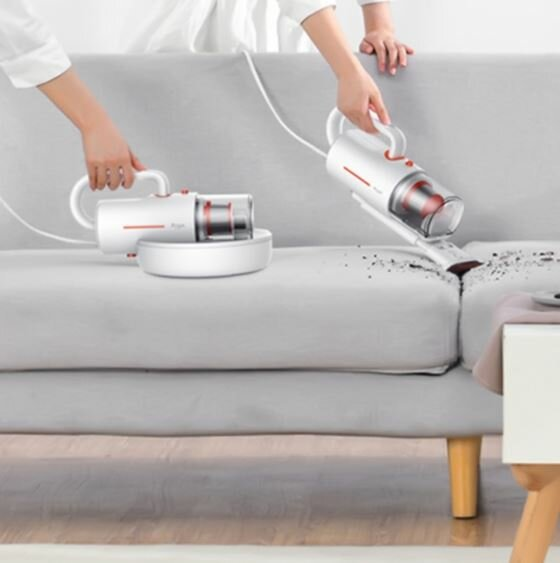 Dwie ręce odkurzają szarą sofę. Jeden odkurzacz spoczywa na podstawie ładującej.