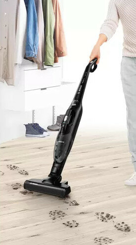 Kobieta używa czarnego odkurzacza, ze śladami butów na drewnianej podłodze. W tle szafa.