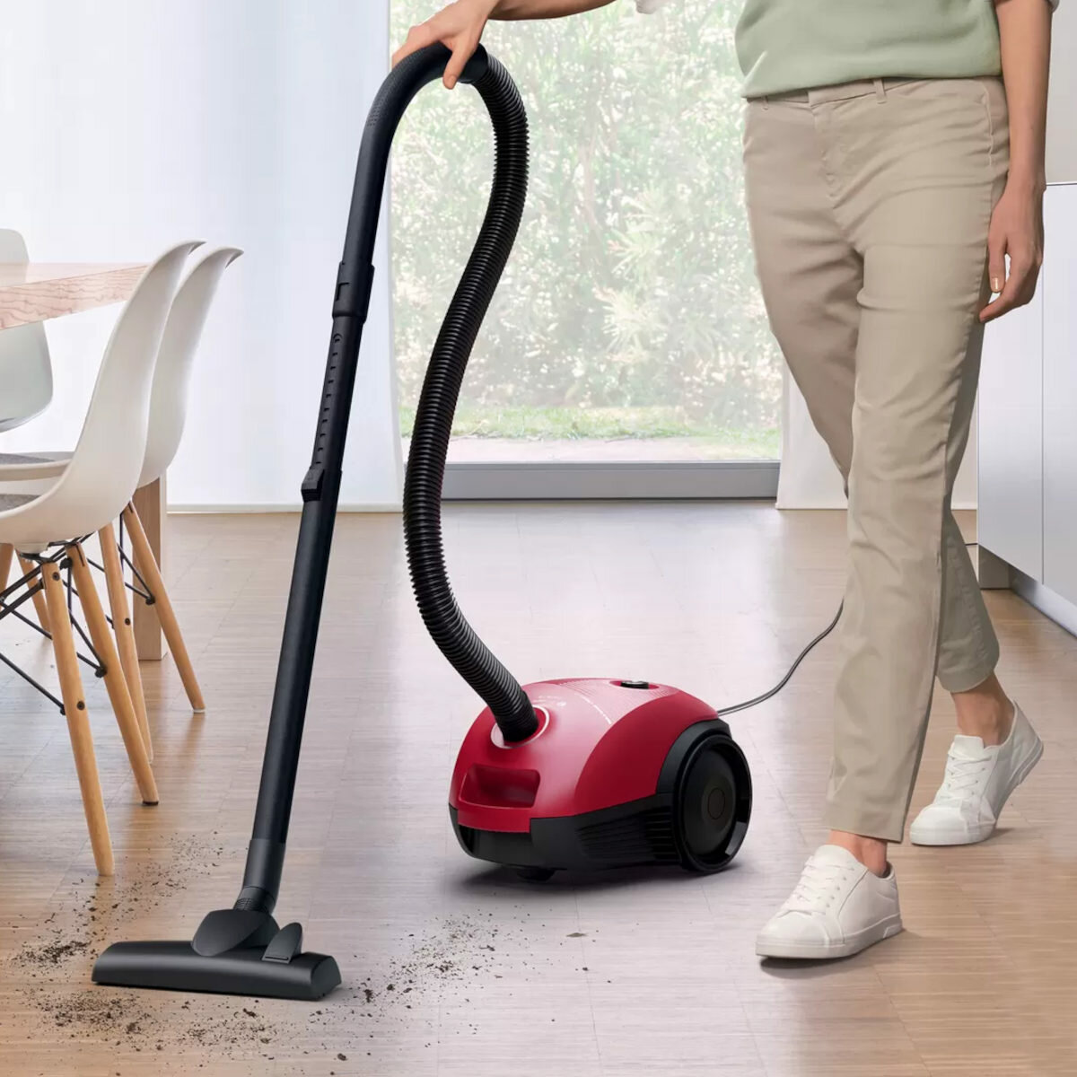 Kobieta odkurza drewnianą podłogę czerwonym odkurzaczem. Są dwa białe krzesła.