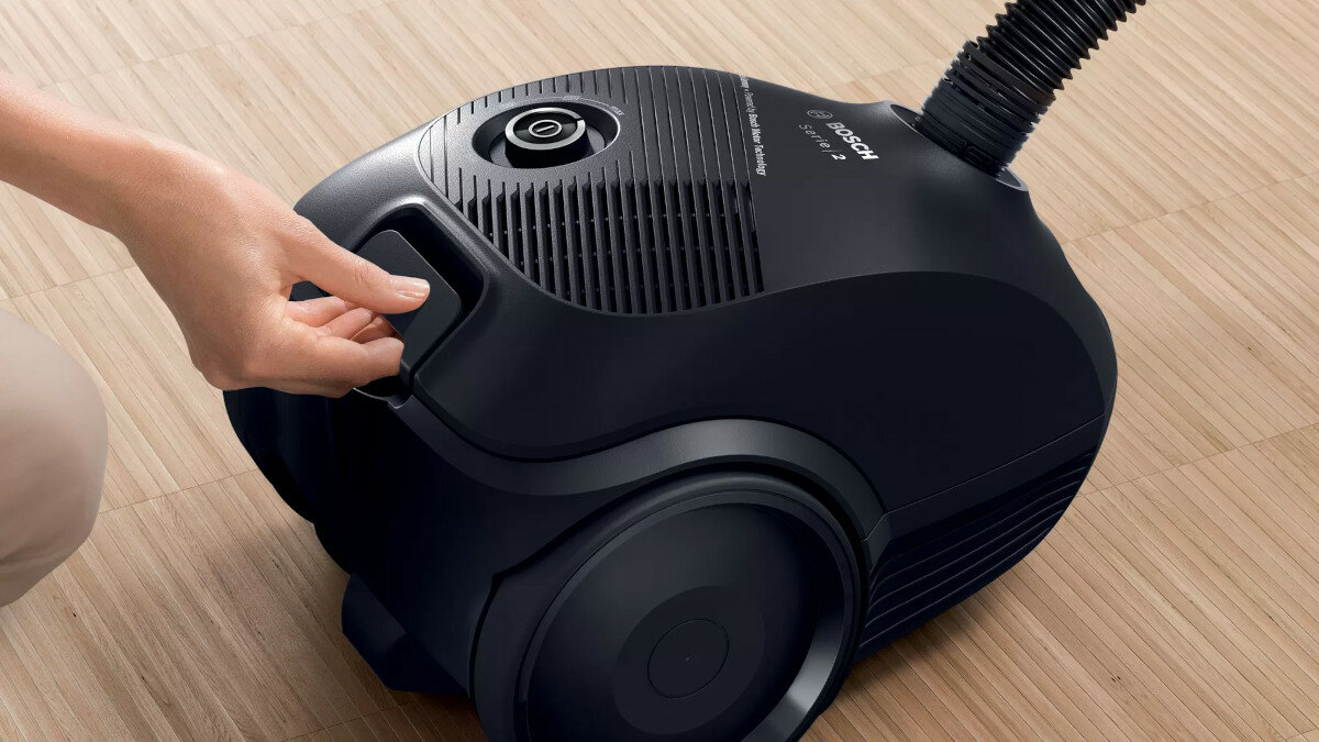 Ręka otwierająca pojemnik na kurz czarnego odkurzacza. Odkurzacz stoi na drewnianej podłodze.