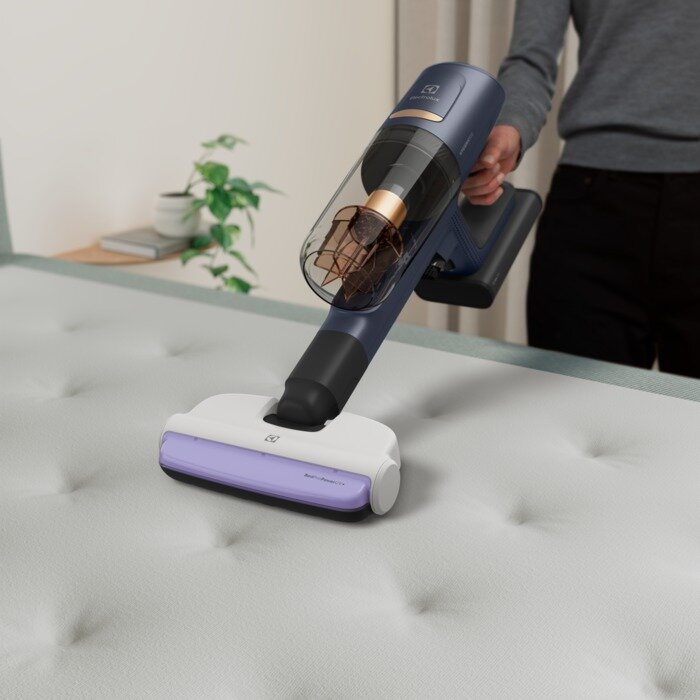 Osoba używa niebieskiego odkurzacza bezprzewodowego na białym materacu.