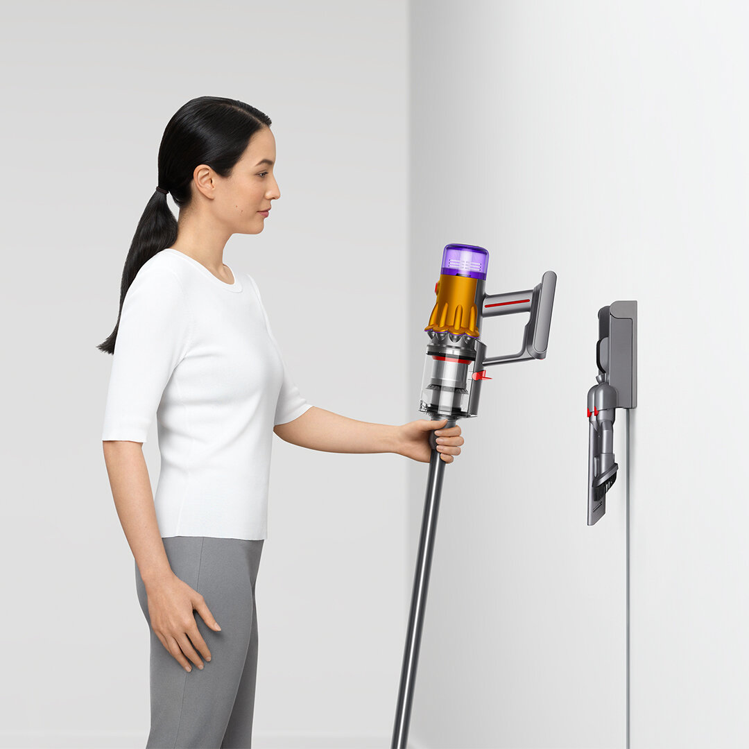 Kobieta trzymająca odkurzacz Dyson obok zamontowanej na ścianie stacji ładowania.