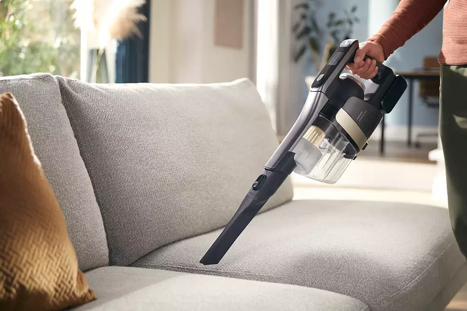 Osoba odkurzająca szarą sofę odkurzaczem ręcznym w nowoczesnym salonie.