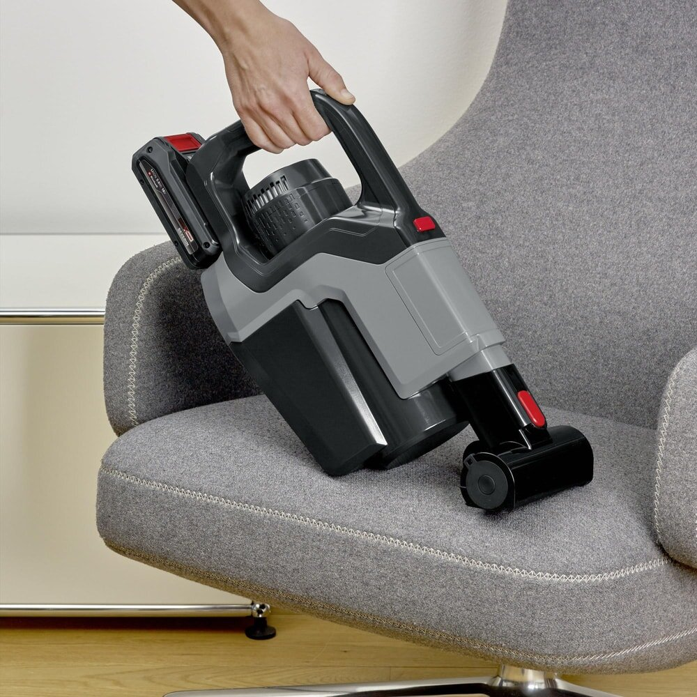 Ręka trzyma szaro-czarny odkurzacz bezprzewodowy nad szarym fotelem.