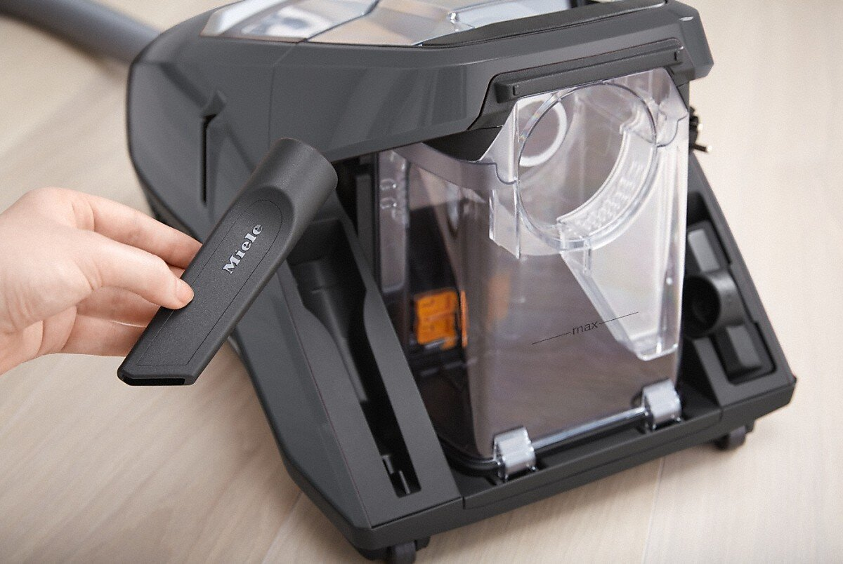 Ręka wyjmuje ssawkę szczelinową z szarego odkurzacza. Widoczne logo Miele.