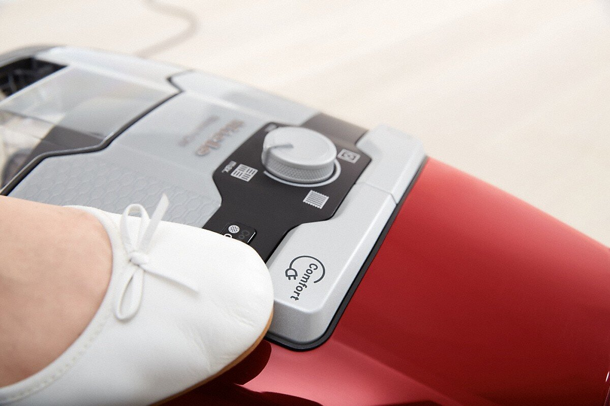 Biały but z kokardą spoczywa na czerwonym odkurzaczu. Odkurzacz ma szare i czarne detale. Widoczne jest pokrętło.