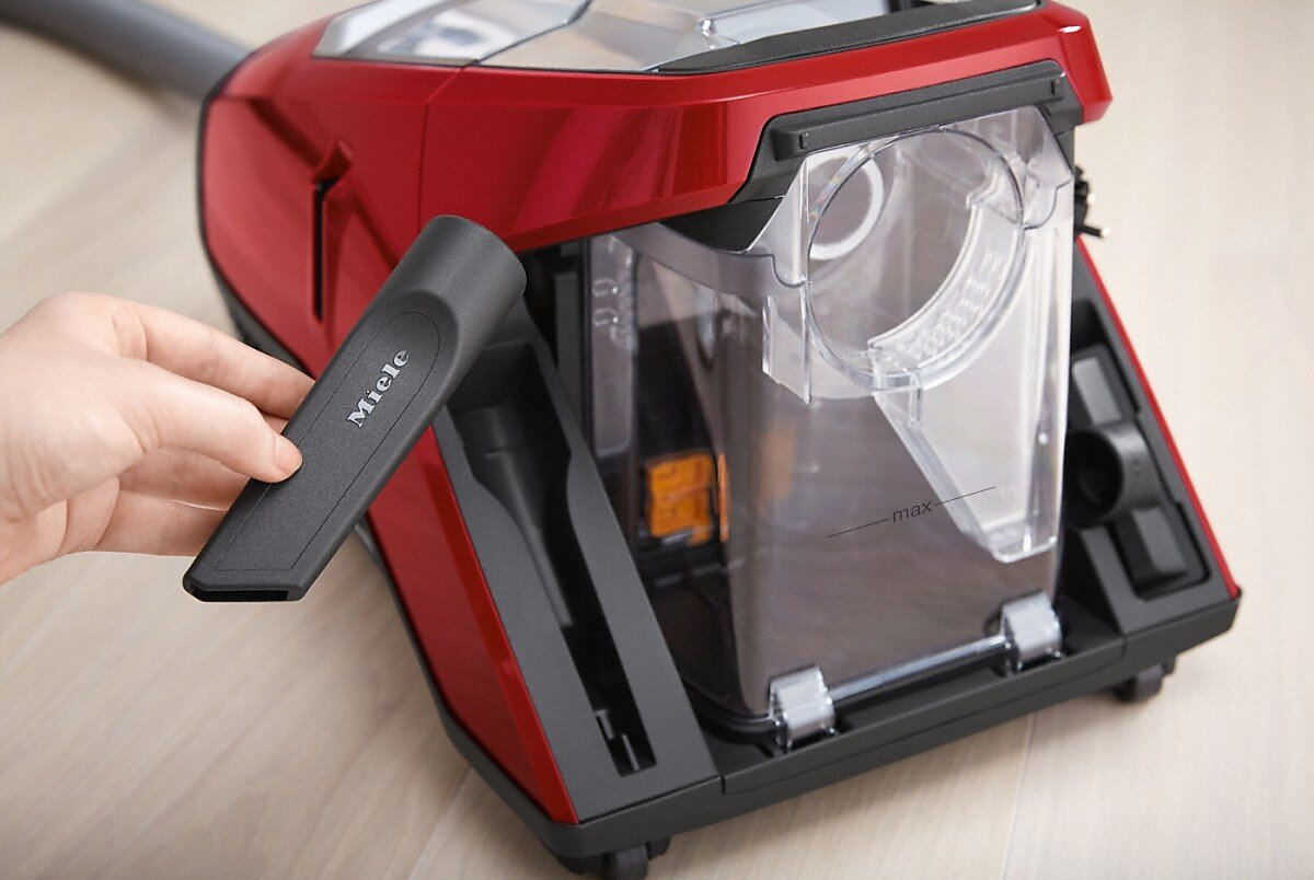 Czerwony odkurzacz Miele z czarną dyszą. Ręka trzyma dyszę w pobliżu przezroczystego pojemnika na kurz.